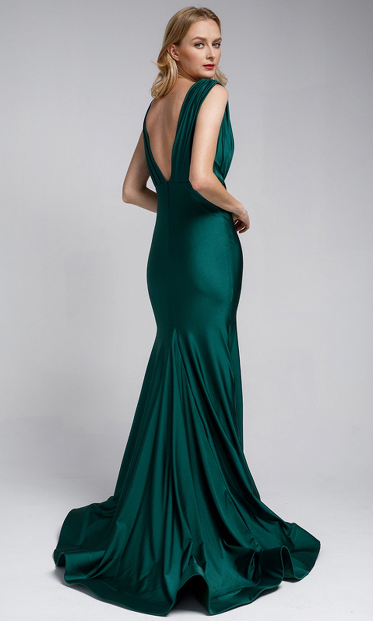 A woman poses against a plain light gray background wearing the Plunging V-Neck Long Lycra Prom Dress: Amelia Couture 370, a floor-length deep green mermaid gown with a low back and slight train.